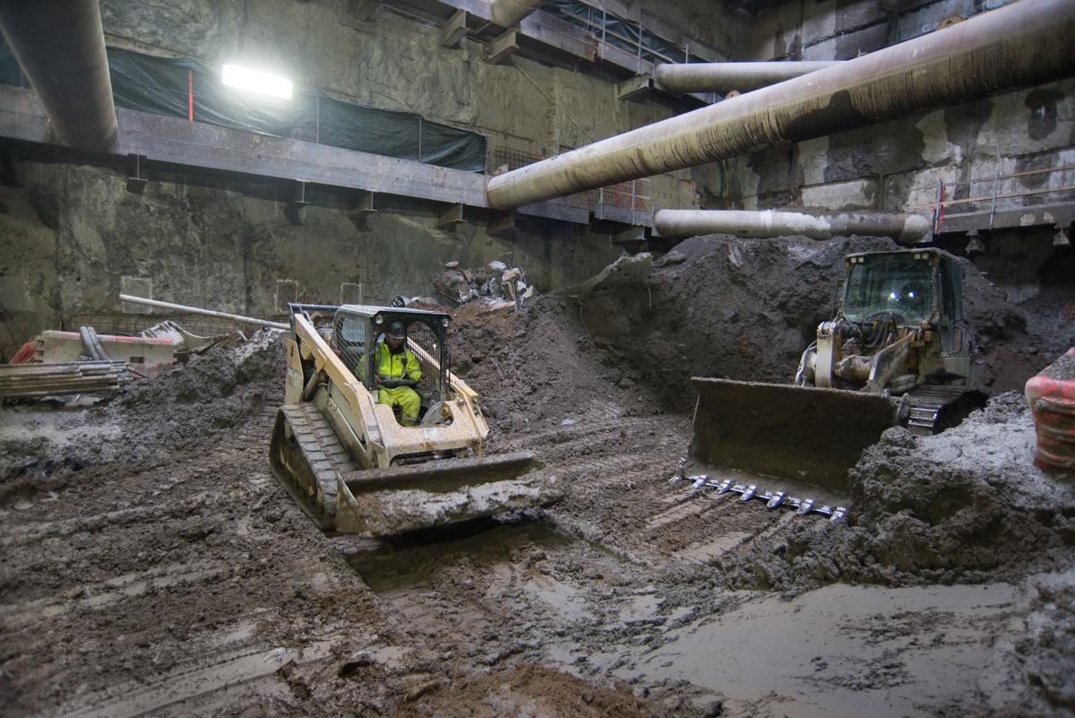 Workers operate heavy equipment to excavate underground station cavern, with large pipes and concrete structure visible above.