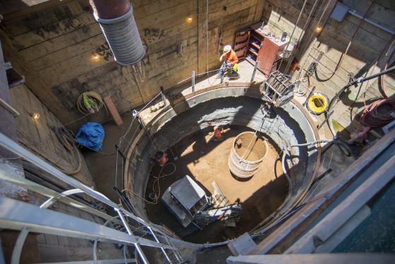 Workers in hard hats conduct underground excavation work in a deep circular shaft with equipment and safety barriers visible.