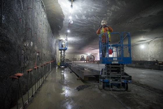 Underground tunnel with wet concrete floor, blue elevated work platform, and construction lighting during sewer installation work.
