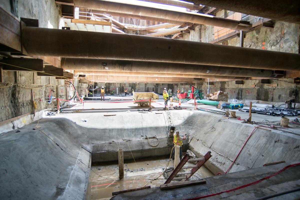 Construction site under highway overpass with concrete pit, rebar, dewatering equipment, and workers installing waterproofing.