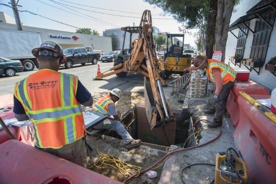 Workers in safety vests excavate street with backhoe for sewer line work on Brannan Street.