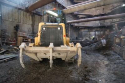 Yellow skid steer loader positioned in excavation site with wooden support beams visible in industrial setting.
