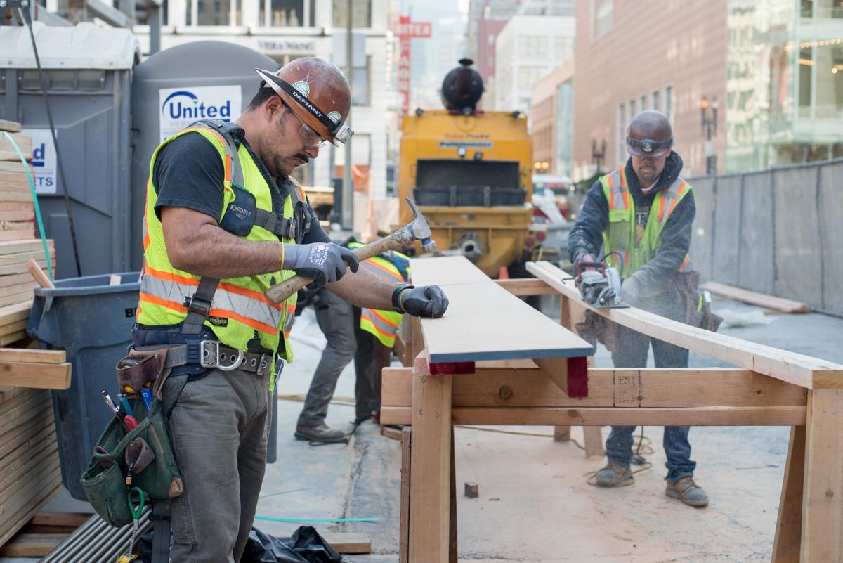 Construction workers in safety gear build wooden formwork for concrete at an urban transit station site.
