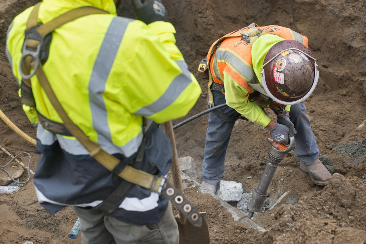 Two construction workers in safety vests and hard hats perform underground utility work in an excavated area.