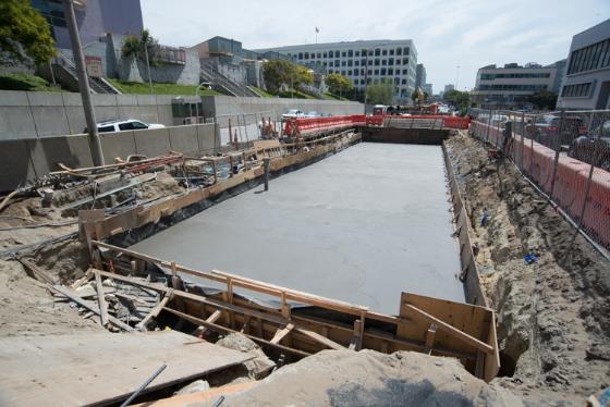 Construction site with concrete foundation poured for Central Subway station, surrounded by steel barriers and urban buildings.