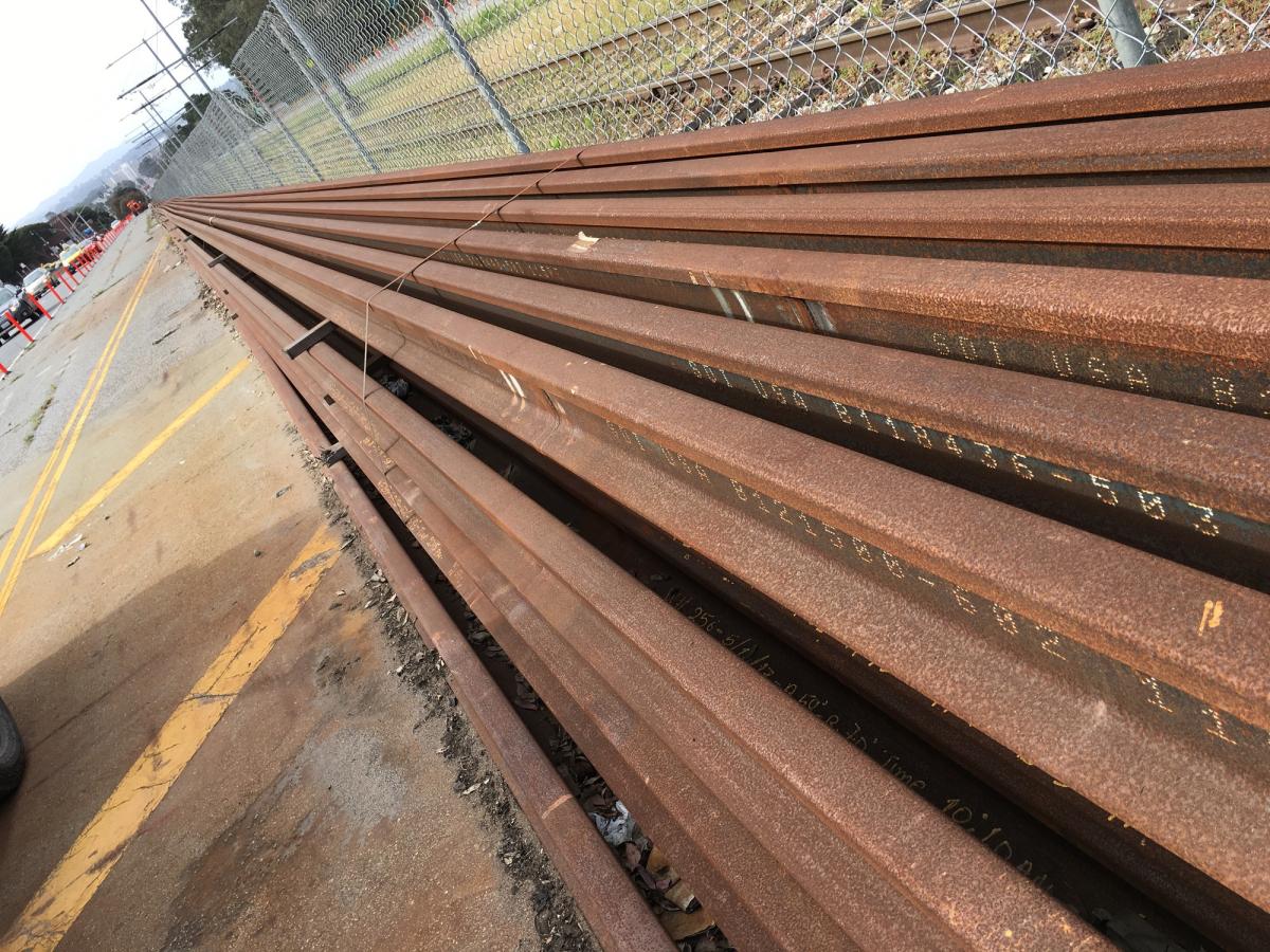 Stacked steel rails lined up along a paved surface next to a chain-link fence.