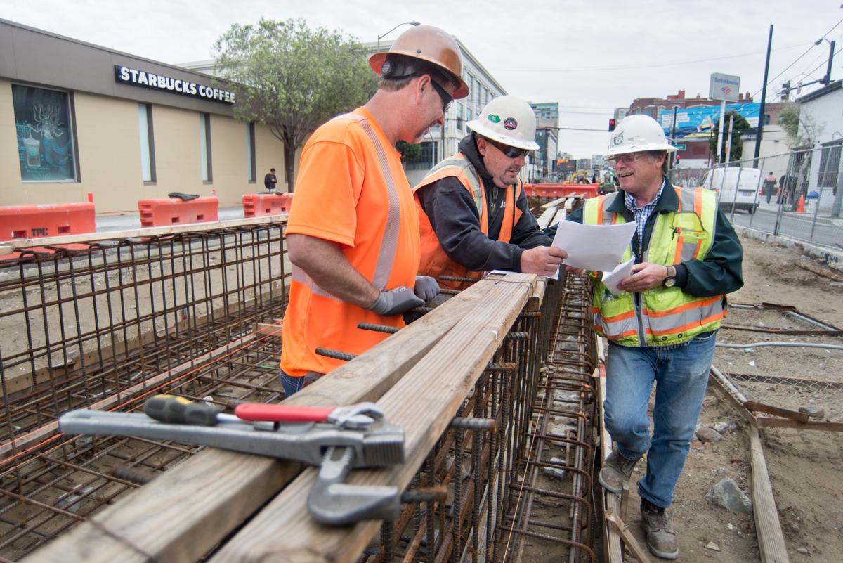 Three construction workers in safety gear review plans at a reinforced concrete platform with rebar framework visible.