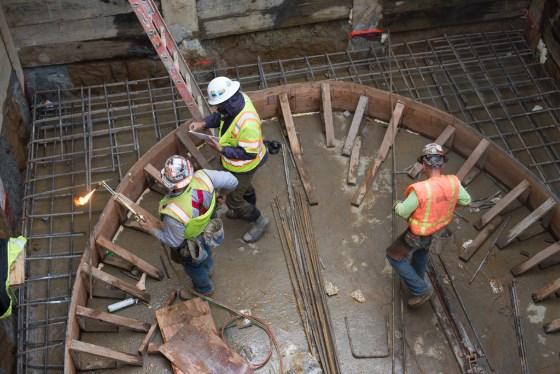 Construction workers in safety vests and hard hats work in an underground shaft with wooden supports and reinforced concrete framework.