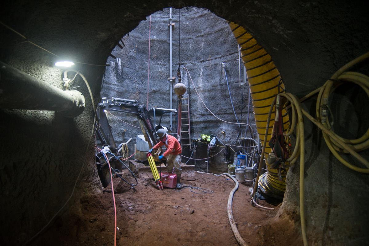 Worker in red jacket prepares equipment at bottom of underground shaft surrounded by coiled yellow cables and metal framework.