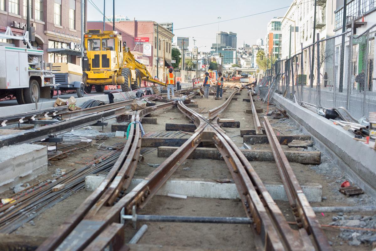 Streetcar track installation work on urban street with yellow construction equipment and workers.