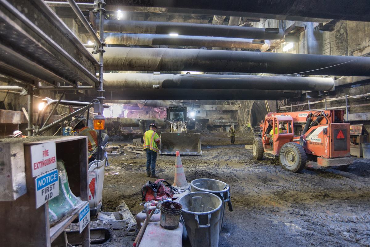 Construction work underway beneath elevated highway structure with orange equipment and workers in safety gear.
