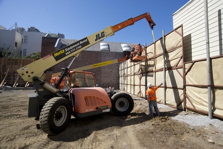 Telescopic handler lifting materials at construction site with worker and sound barrier wall.