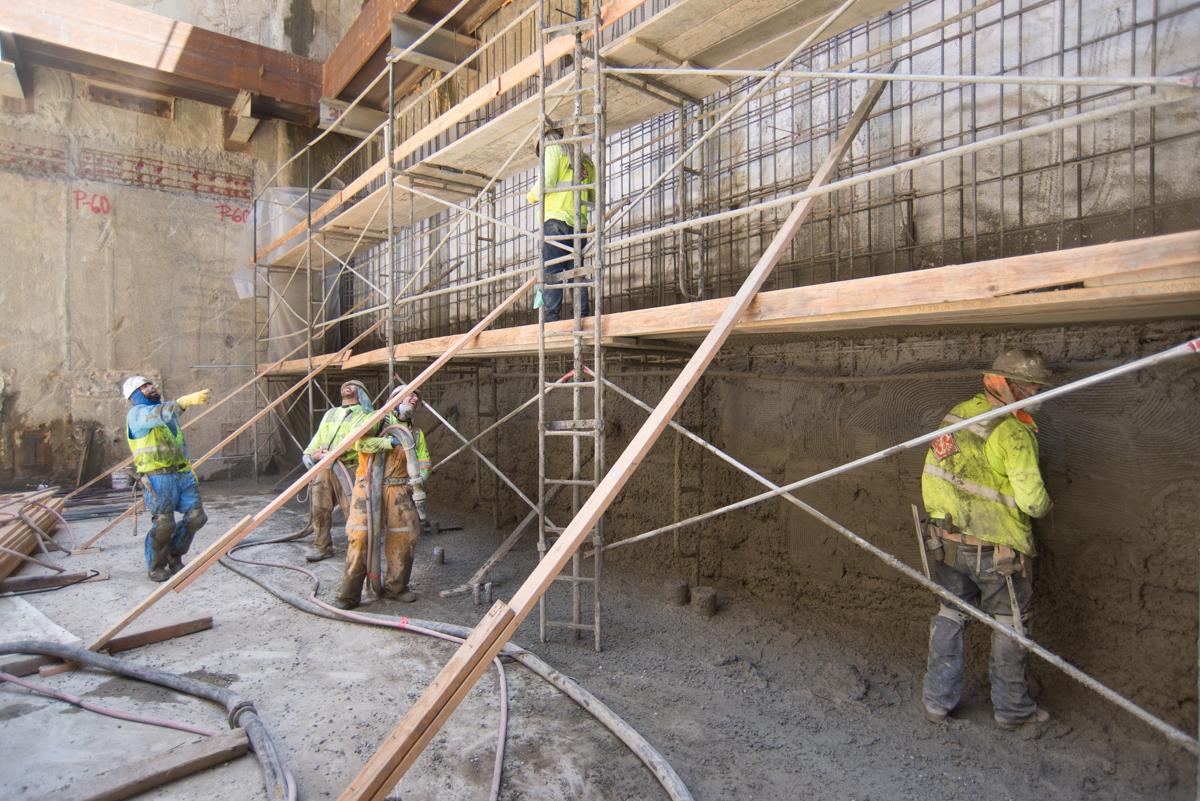 Construction workers in safety vests on scaffolding inside a concrete transit station structure under renovation.