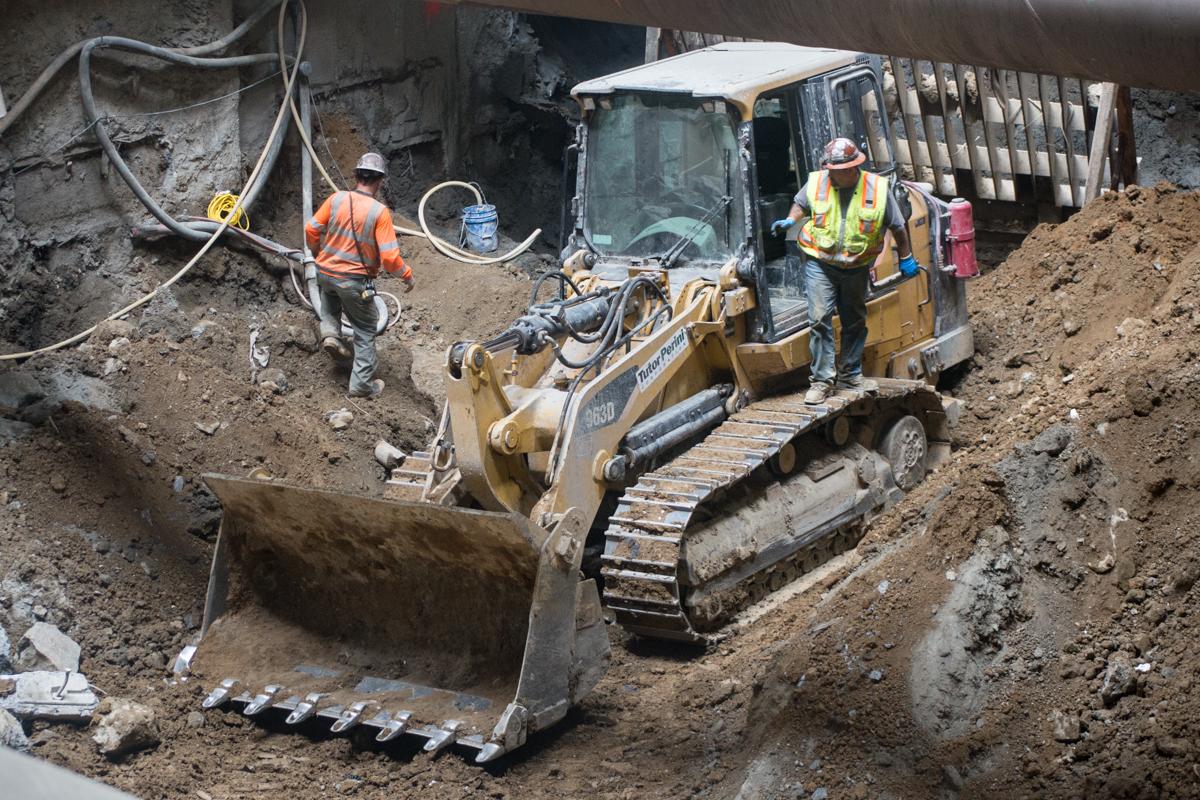 Construction workers operate an excavator to remove soil and rock from an underground transit station excavation site.