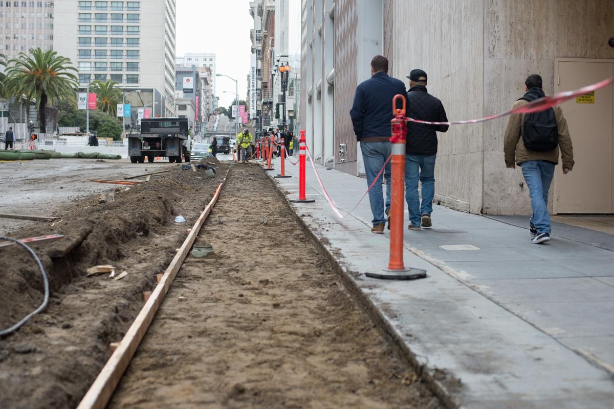 Urban street with construction trench, orange safety barriers, and pedestrians on sidewalk near tall buildings.