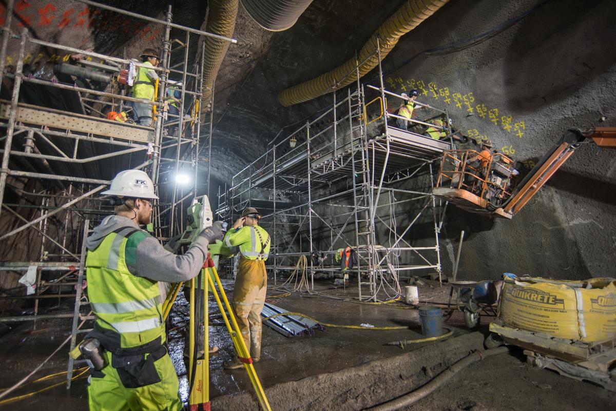 Workers in safety gear operate surveying equipment on scaffolding inside an underground transit tunnel during construction.