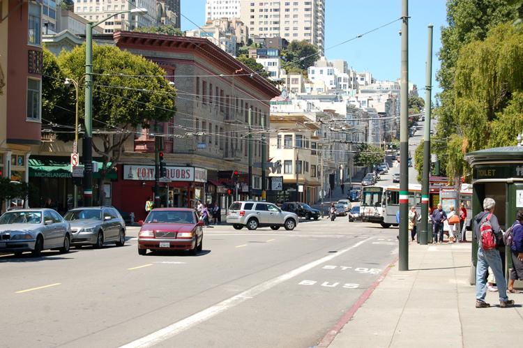 Stockton Street in Chinatown with parked cars, storefronts, bus stop, and residential buildings on hillside.