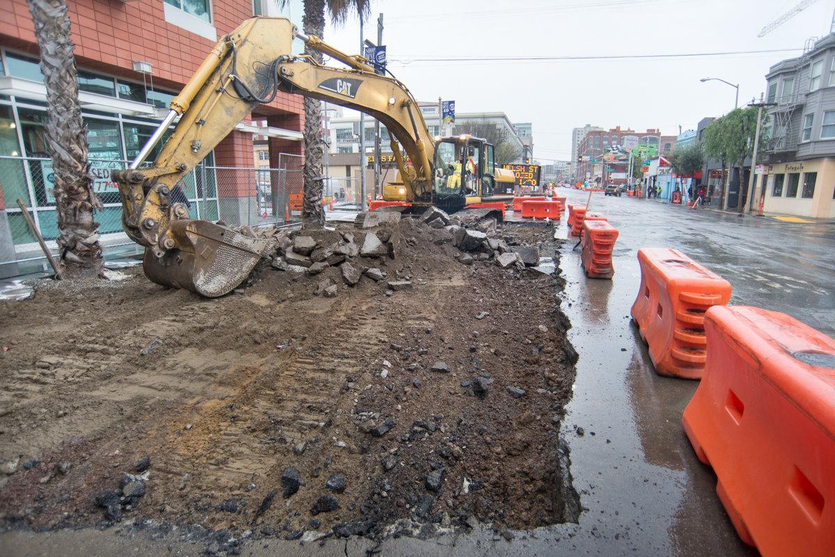 Excavator removing pavement on 4th Street for utility work, with orange traffic barriers lining the wet street.