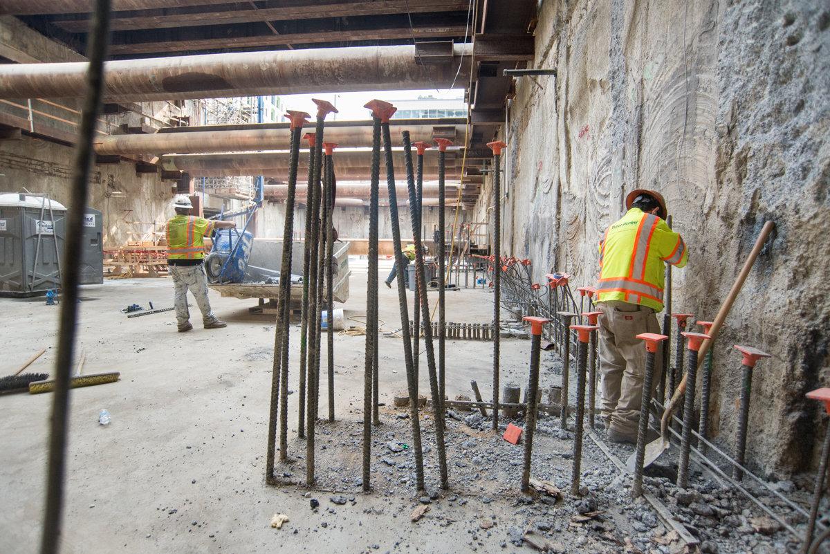 Construction workers in safety vests work on rebar installation along a concrete wall at a transit station headhouse.