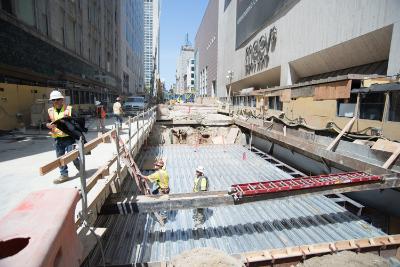 Construction site at Geary and Stockton with workers, equipment, and exposed foundation work on urban street.