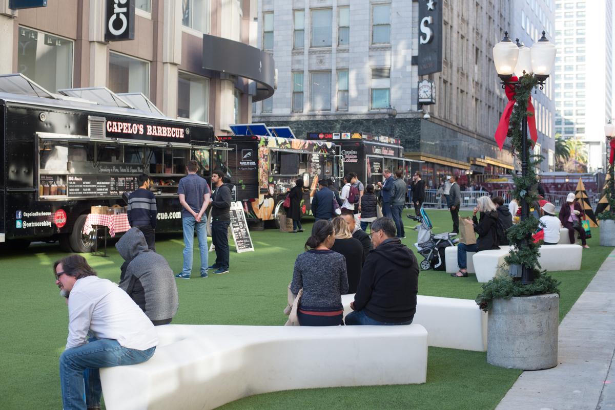 Outdoor urban plaza with food trucks, white curved seating, holiday decorations, and pedestrians on artificial turf.