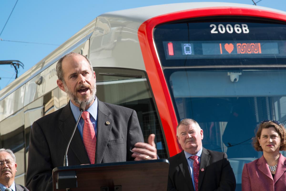 Man in suit speaking at podium in front of red and white light rail vehicle 2006B with officials present.