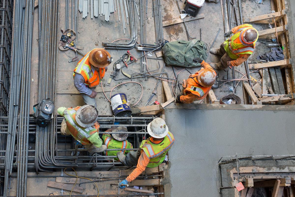 Construction workers in safety gear install rebar and structural elements at underground transit station site.