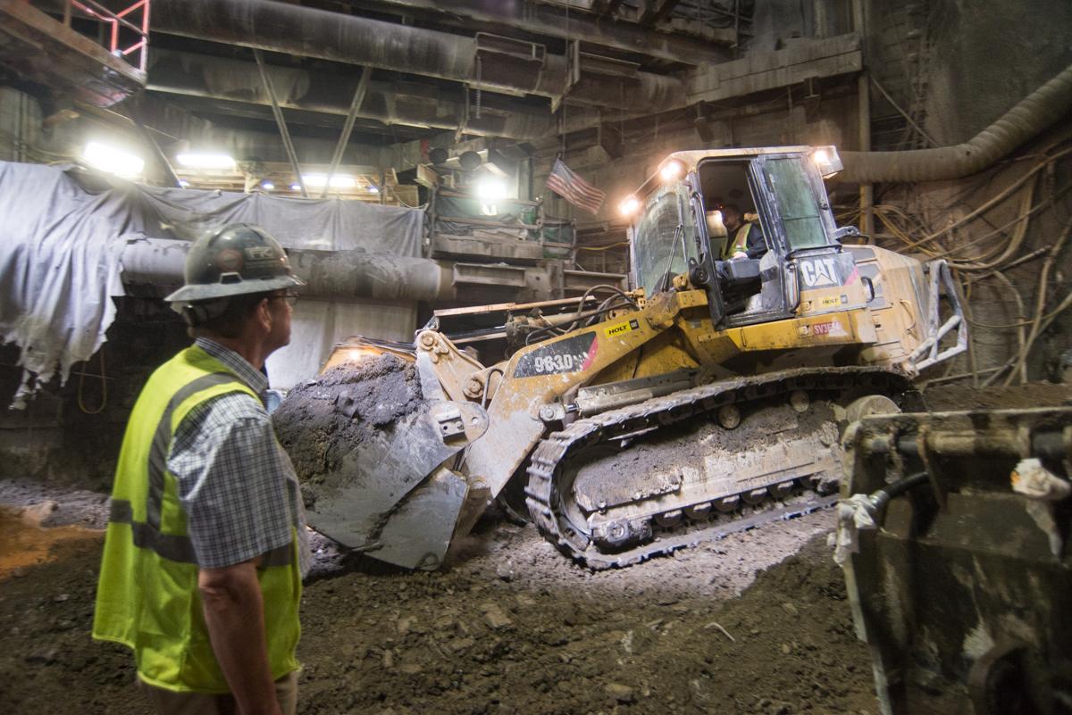 Worker in safety gear supervises yellow excavator loading debris in underground tunnel with industrial lighting.