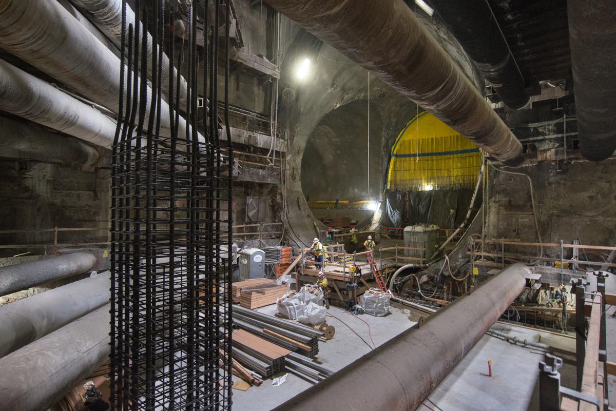 Underground tunnel construction site with workers, reinforced steel rebar framework, and large yellow equipment under bright lighting.