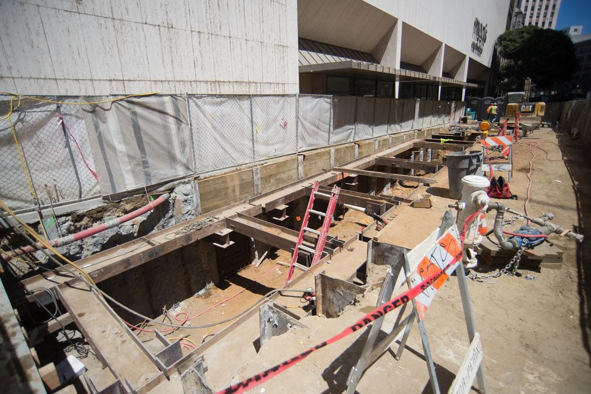 Construction site showing excavated station area with temporary bracing, pink safety barriers, and steel beams under Stockton Street.