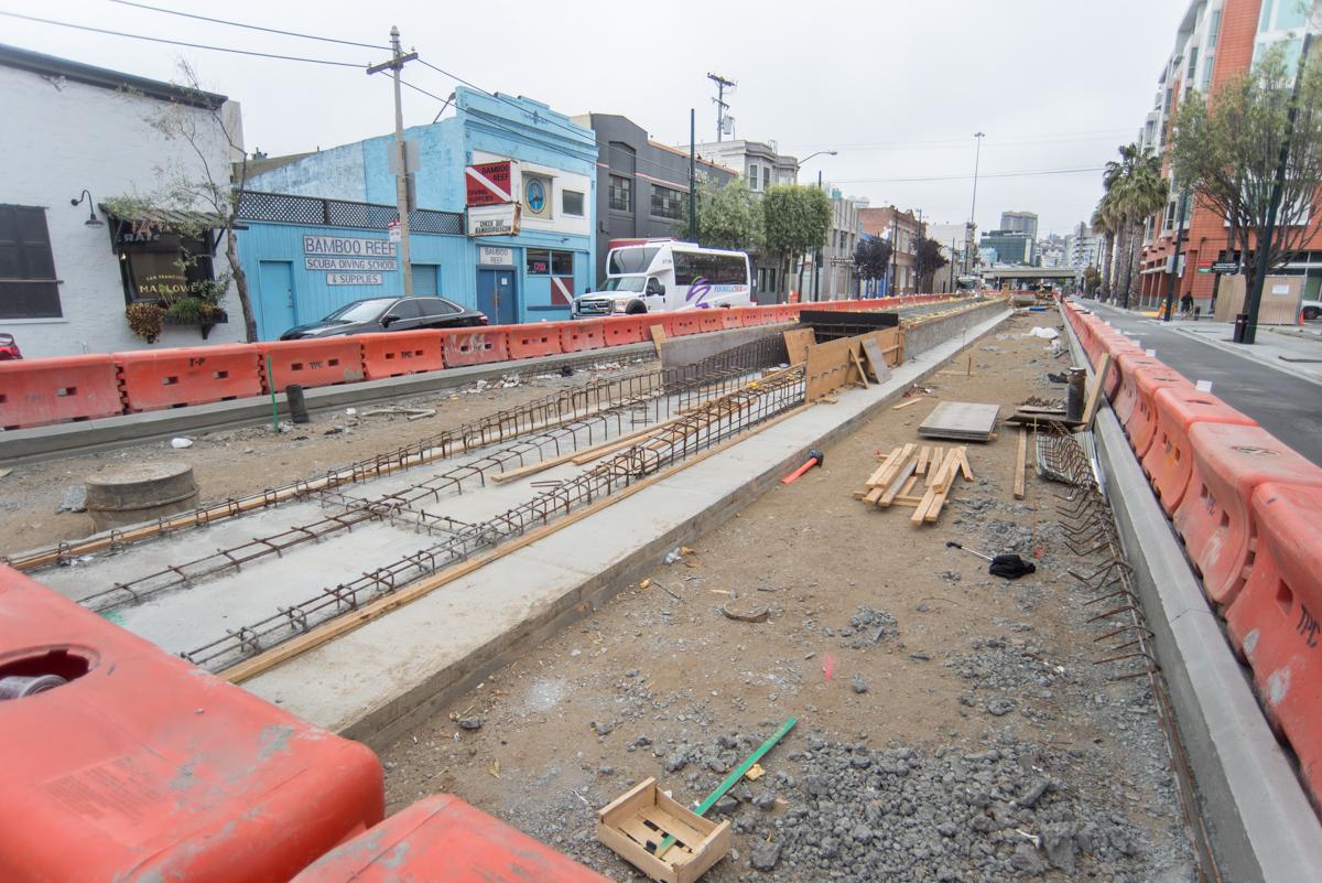 Construction site showing excavated trench with reinforcing steel and concrete foundation work for transit station in urban area.