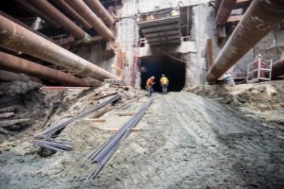 Workers in hard hats stand in tunnel excavation beneath steel support beams and large pipes on 4th Street.