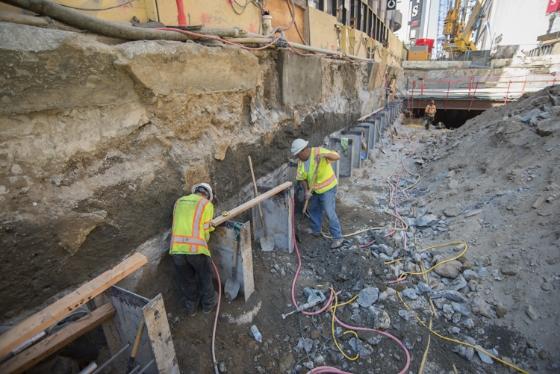 Two workers in safety vests remove concrete around steel I-beams at an urban construction site.