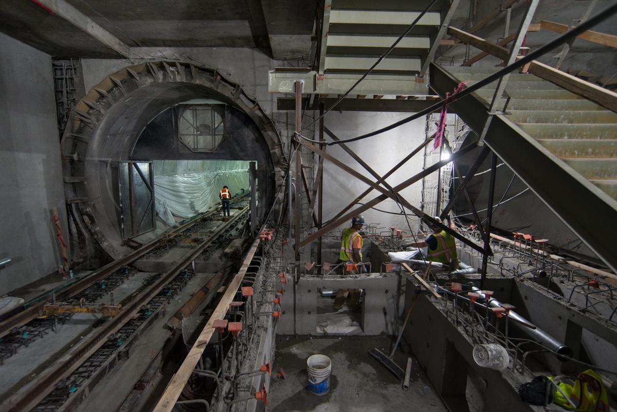 Tunnel boring machine and construction workers inside underground transit station excavation with rail tracks and steel framework visible.