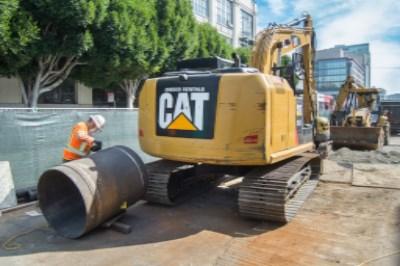 Yellow CAT excavator with worker positioning large sewer pipe section during underground utility construction.