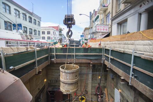 Construction site with steel bucket being lowered into underground shaft for soil removal during transit station excavation.