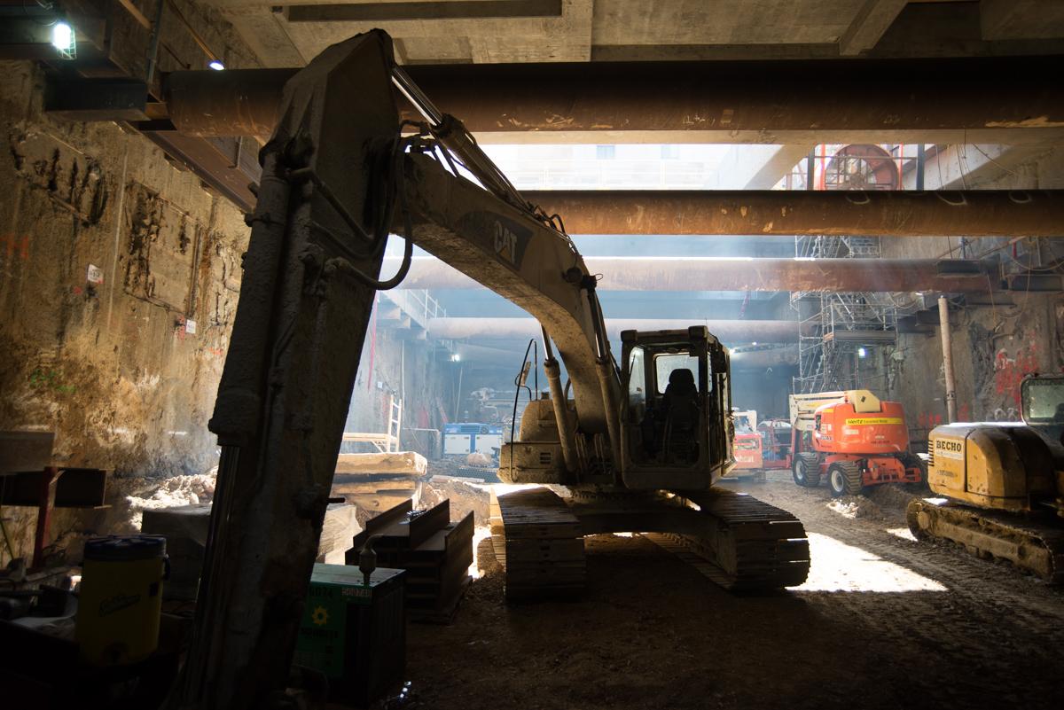 Excavator inside underground construction headhouse with concrete walls and steel support beams visible.