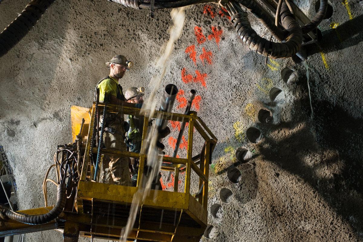 Worker in yellow safety vest on elevated platform operating equipment near concrete wall with red markings and pilot holes.