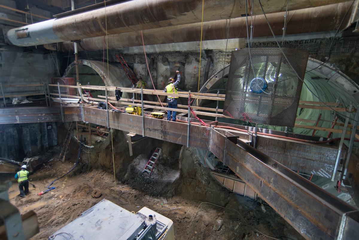 Workers in safety vests install temporary bracing at transit station construction site with excavation underway below.