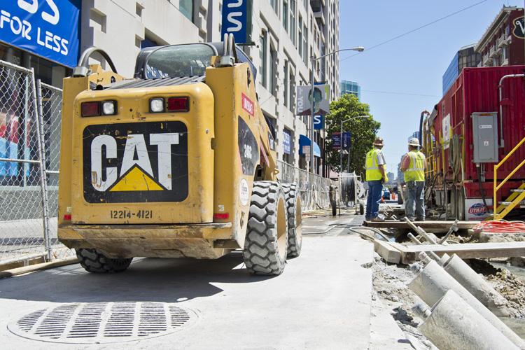 Yellow CAT skid steer loader on urban street with construction workers and equipment during daytime roadwork.