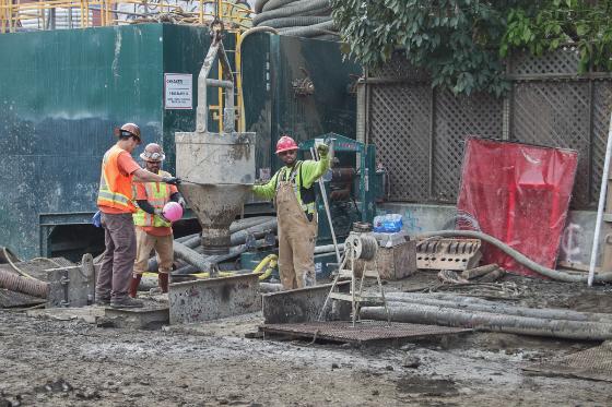 Construction workers in safety vests operate equipment at a job site on a city street, with concrete pipes and materials visible nearby.