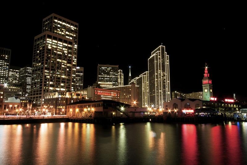 San Francisco Embarcadero waterfront at night with illuminated skyscrapers, Ferry Building clock tower, and holiday lights reflecting on water.