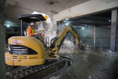 Worker operating yellow excavator inside underground garage during construction and demolition work.