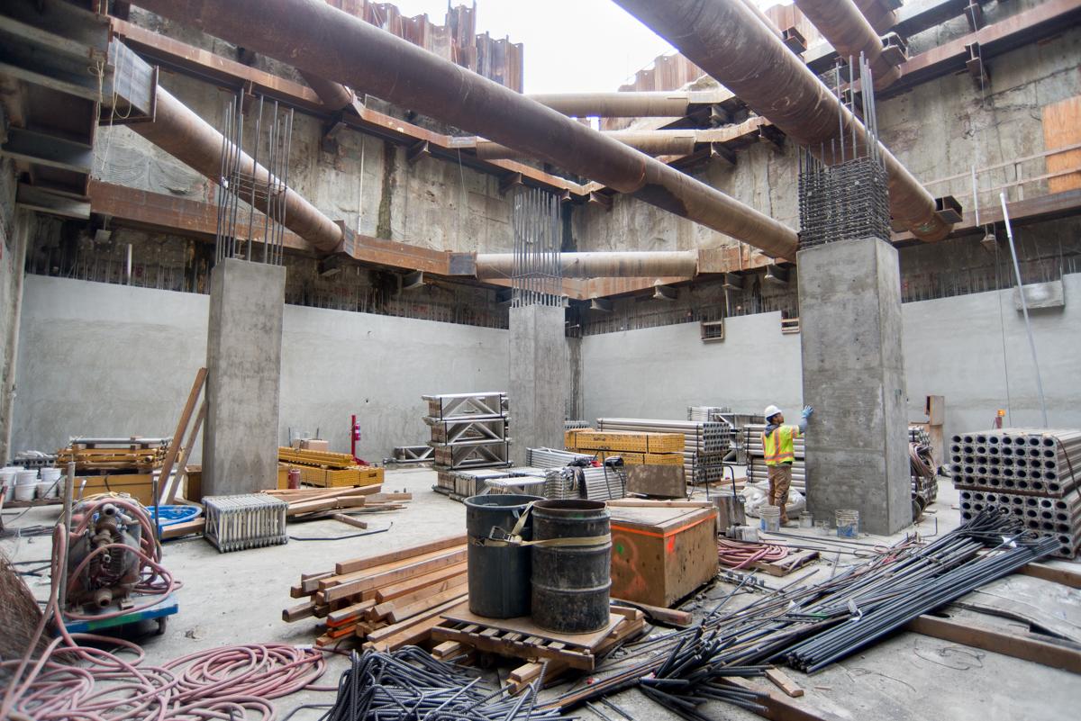 Interior construction of transit station with concrete columns, steel beams, building materials, and workers in progress.