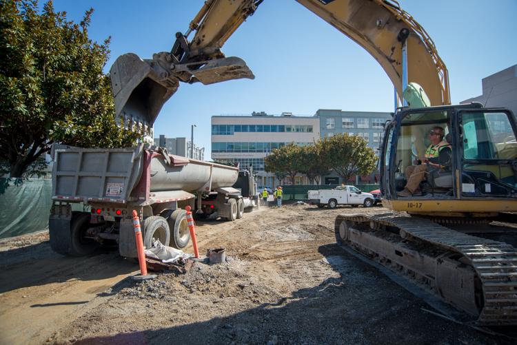 Excavator loading debris into dump truck at urban construction site with utility work underway.