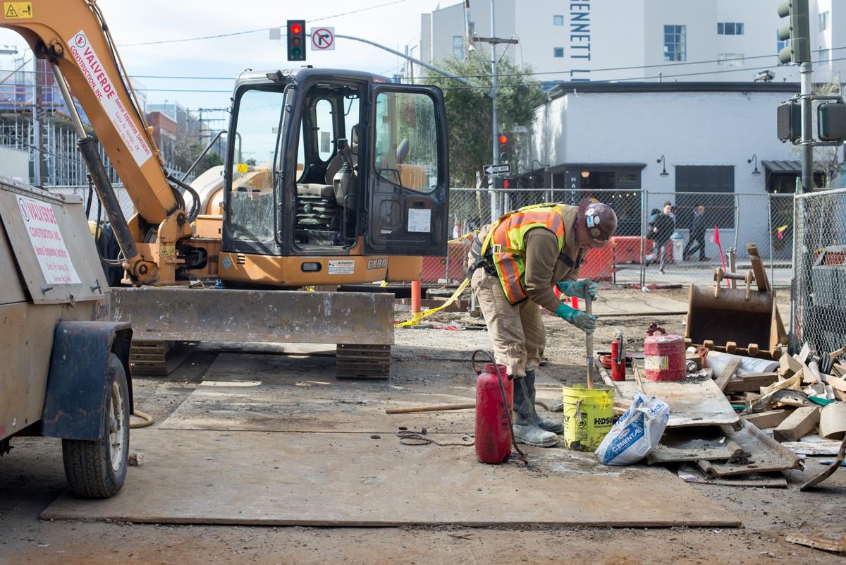 Construction worker in safety vest operating equipment near excavator on 4th Street utility trench work site.