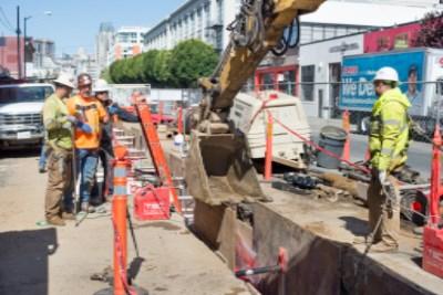 Construction workers lower pipe into trench on 4th Street for utility installation work.