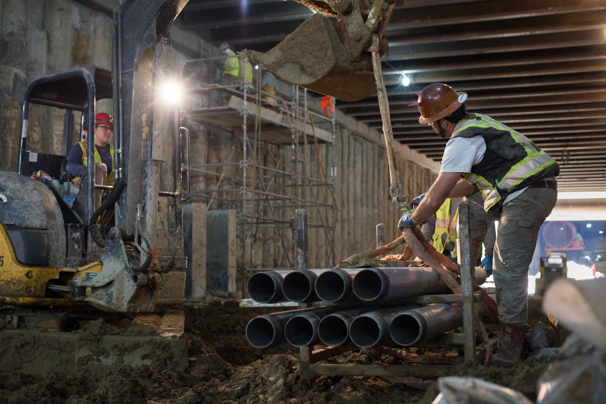 Workers unload large metal pipe sections at an underground transit station construction site.