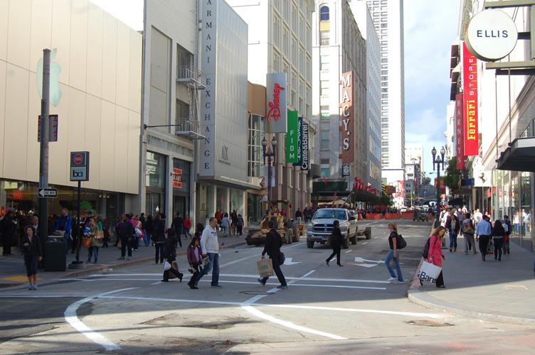 Busy downtown street intersection with pedestrians crossing, storefronts, and tall buildings lining both sides.