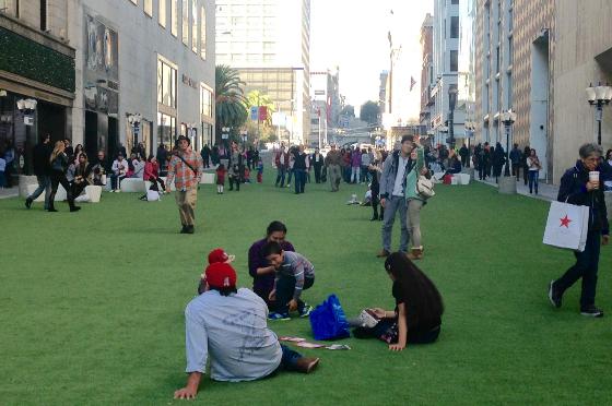 People relaxing on artificial grass in an urban plaza surrounded by tall buildings and shops.